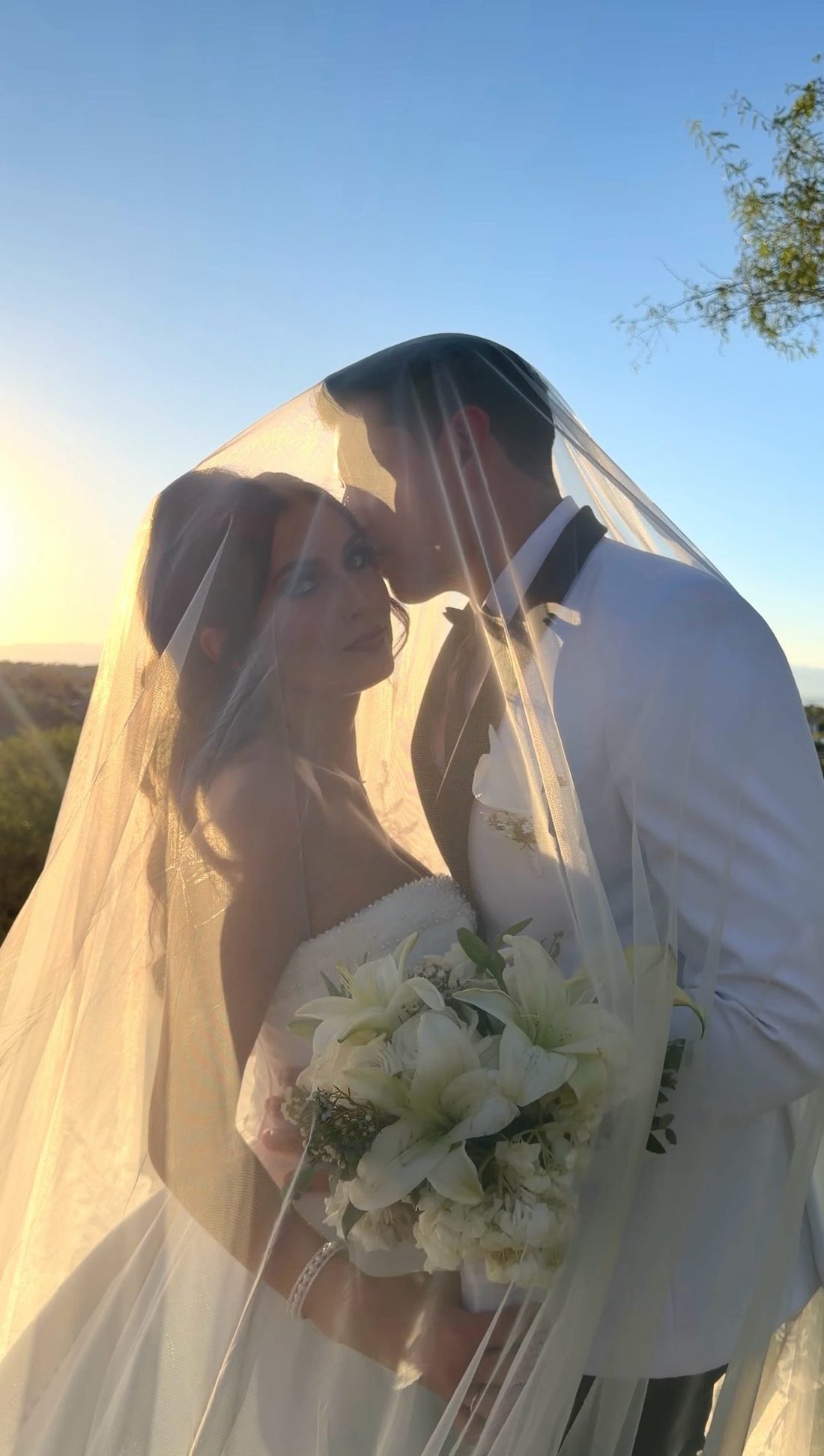 Groom kissing the bride on the forehead beneath her veil at sunset in Las Vegas, the bride holding a white lily bouquet