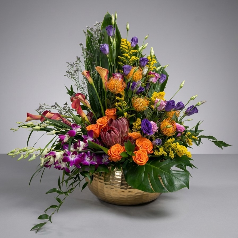 Vibrant tropical floral centerpiece with orange roses, purple orchids, pincushion proteas, and a large monstera leaf in a hammered gold bowl.