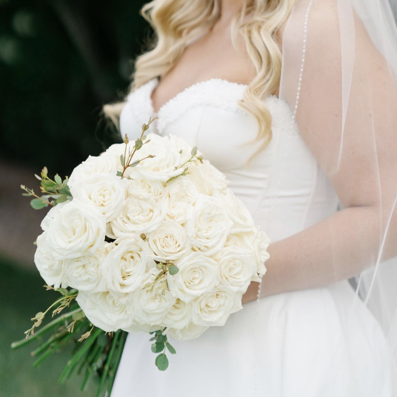 Bride holding a large round wedding bouquet of white roses with subtle eucalyptus
greenery.